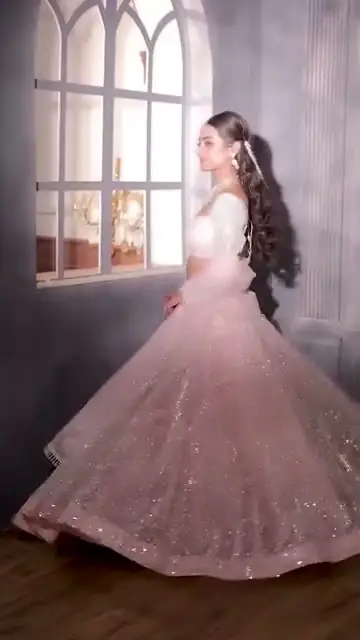 A bride standing facing the camera, hands folded at her waist, wearing a pale pink sequinned lehenga with a beaded ivory blouse and a layered emerald necklace.